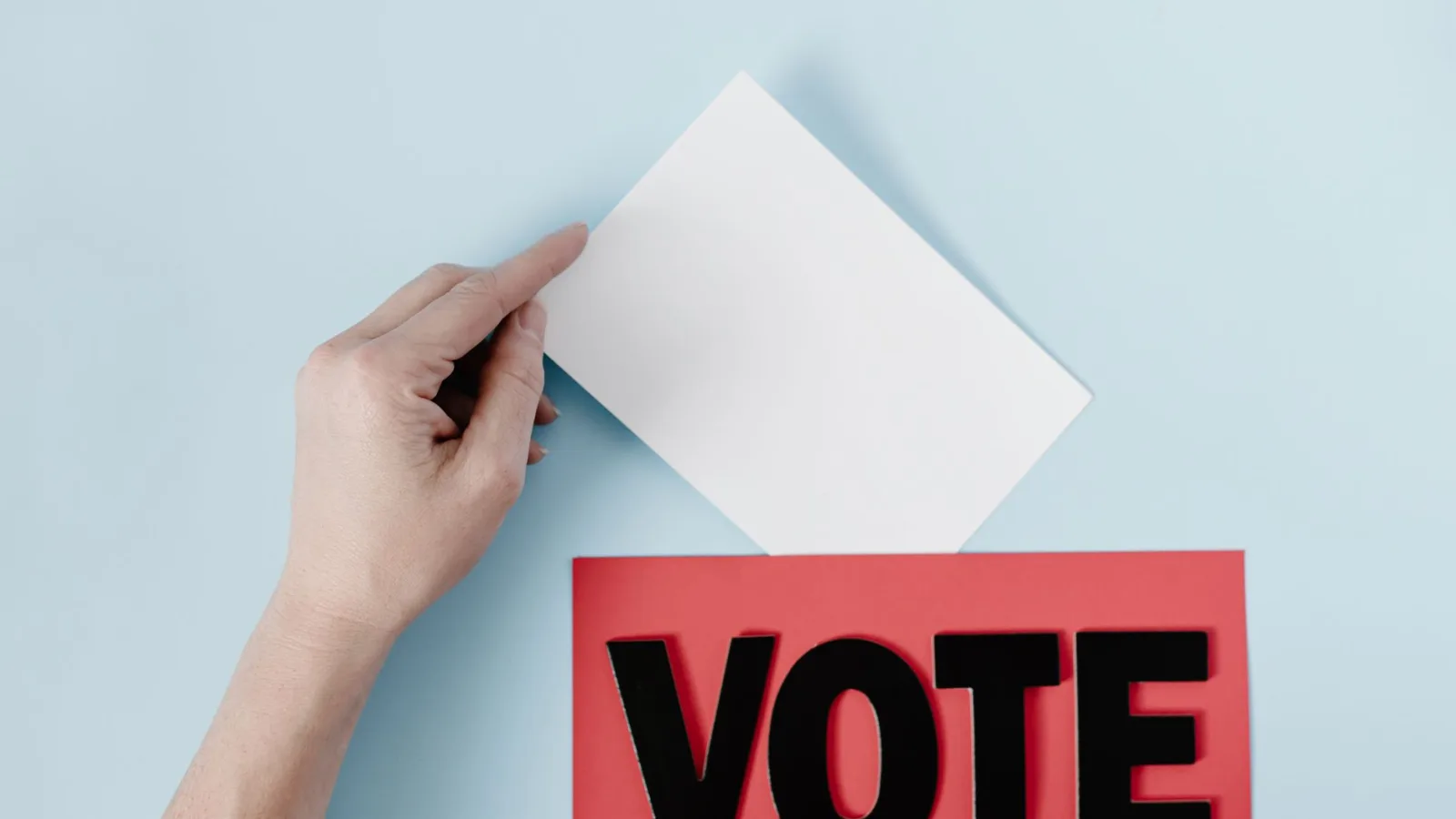 A hand places a ballot into a red box with the word VOTE against a blue background.