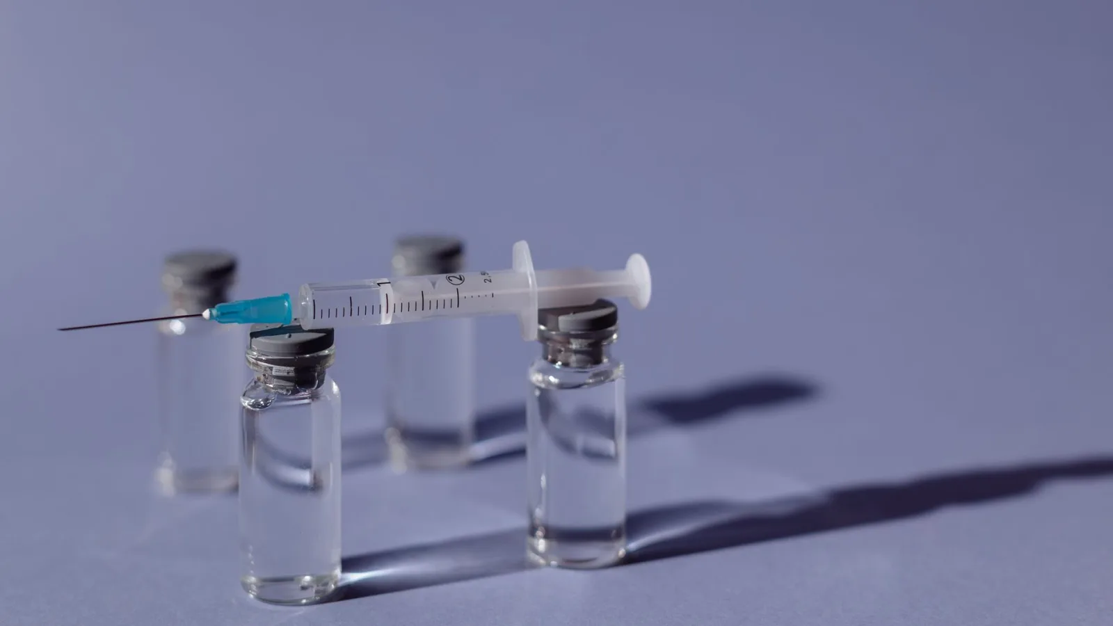 A syringe balanced on vaccine vials with a purple backdrop, representing medical treatment.